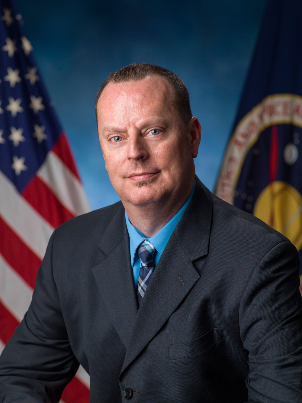 Professional portrait of a man wearing a navy blue suit, light blue shirt, and a blue patterned tie.